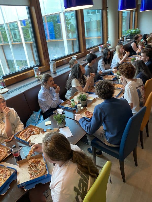 Leerlingen eten pizza en praten met elkaar aan een lange tafel bij het raam